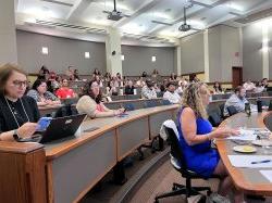 Attendees are seated at long rounded tables in a classroom.